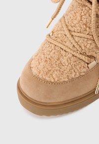 Ruskean mokkanahka- ja teksturoitu fleece-saapas, jossa on punottu nauha ja kultasävyiset yksityiskohdat. Pyöreä kärki ja kumipohja, jossa on pieni alusta.