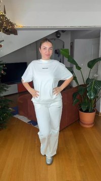 Hvit bomulls-T-skjorte med rund hals og logo på brystet, kombinert med matchende hvite bukser. Jevn tekstur, satt i et lyst interiør.