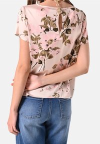 Blouse florale rose clair à manches courtes, avec un détail dos en forme de trou de serrure. Associée à un jean bleu en denim, affichant un style décontracté.