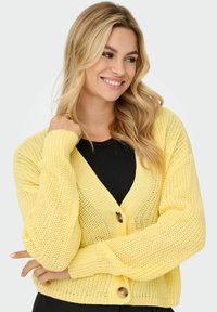 Dzeltena adīta kardigāna ar V veida kakla izgriezumu un pogu aizdari. Iezīmējas ar plašām, teksturētām piedurknēm un brīvu piegriezumu, izgatavota no viegla materiāla.
