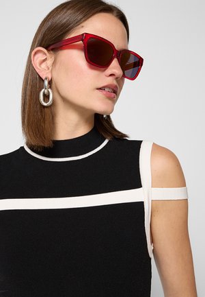 Donna con capelli castani lisci che indossa occhiali da sole rossi quadrati, grandi orecchini a catena in argento e una canotta nera con strisce bianche.