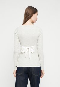 Ανοιχτό γκρι μπλούζα μακρυμάνικη με σχέδιο πεπλούμ και λευκή κορδέλα δέσιμο στο πίσω μέρος, συνδυασμένη με σκούρα μπλε τζιν. Λείο υφασμάτινο ύφασμα.