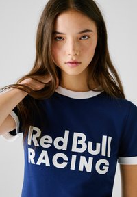 Giovane donna con lunghi capelli castani, che indossa una maglietta blu navy con bordi bianchi e la scritta "Red Bull Racing" stampata sul davanti, all'interno.