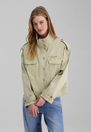Young woman with long hair wearing a light beige buttoned jacket and blue jeans, standing against a plain gray background.