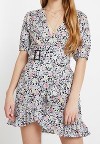 Vestido floral con mangas abullonadas, hebilla en la cintura, dobladillo con volantes y collares en capas en mujer con cabello largo.