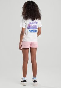 White cotton t-shirt with colorful graphic on back, paired with light pink denim shorts. Model wearing white sneakers with blue and white striped socks.