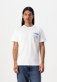 T-shirt en coton blanc avec un col rond, arborant un texte graphique bleu "TOMMY" et un design coloré sur la partie gauche de la poitrine. Jean en denim bleu.