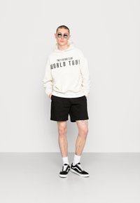 Cream hoodie with "WORLD TOUR" text, black shorts, black Vans shoes, and sunglasses. Model stands against a plain white background.