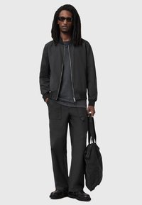 Czarna kurtka bomberka z zamkiem, szary T-shirt, luźne czarne spodnie cargo i czarne buty. Trzyma ciemną torbę na ramię.