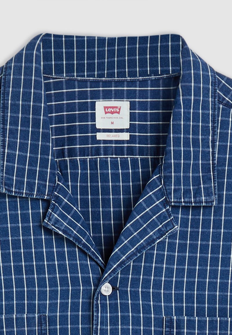 Camisa de algodón a cuadros azules con cuello en pico, que presenta rayas verticales y horizontales blancas, y un cierre con un solo botón.