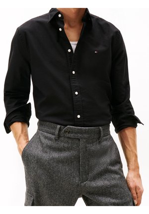 Homme portant une chemise noire à boutons avec les manches retroussées et un pantalon gris à motif chevrons, les mains dans les poches.