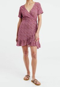 Vestido envolvente floral rosa con mangas cortas, dobladillo con volantes y detalle de lazo en la cintura. Tejido ligero y texturizado con un patrón juguetón.