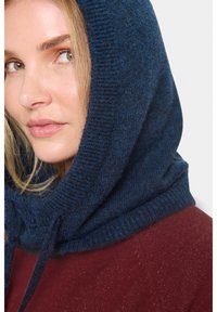 Žena so svetlou pokožkou a zelenými očami, ktorá má na sebe modrý pletený kapucňový sveter a červený textúrovaný sveter, pozerá sa bokom proti jednoduchému pozadiu.