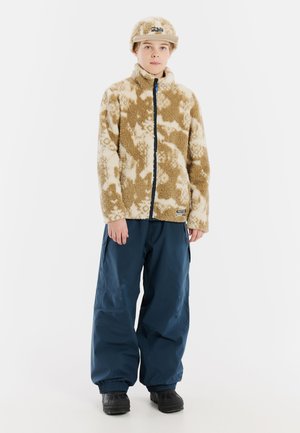 Fleece jack in tan met crème patronen, gecombineerd met marineblauwe broek. De outfit heeft een strakke pet en stevige zwarte laarzen.