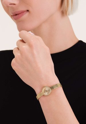 Reloj de malla dorada con una esfera redonda, esfera plateada y acentos de cristales. Se lleva en la muñeca, mostrando una banda de metal texturizada.