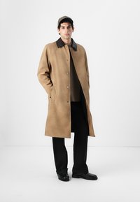 Manteau en suède beige avec un col noir, des boutons à l'avant et des poches latérales. Le modèle porte un haut sombre, un pantalon noir et une casquette à carreaux.