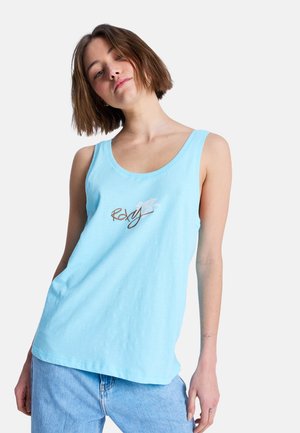 Jeune femme aux cheveux bruns courts portant un haut sans manches bleu clair avec le logo "Roxy" et un jean bleu clair, debout devant un fond uni.