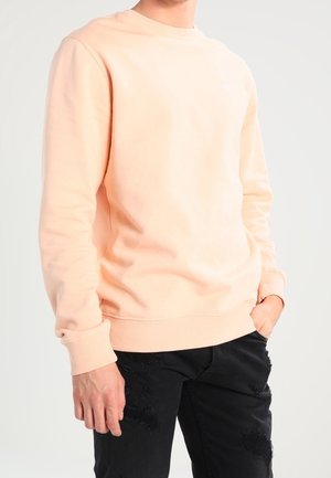 Persona con un sudadera de manga larga en melocotón claro y jeans negros desgastados, con una mano en el bolsillo contra un fondo sencillo.