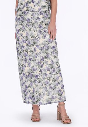 Donna che indossa una lunga gonna floreale con fiori viola e grigi, abbinata a sandali beige con tacco a blocco e punta aperta, in piedi davanti a uno sfondo bianco.