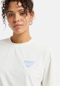Camiseta blanca de manga corta hecha de tela suave, con un logo azul claro "ROXY ACTIVE BY NATURE" en el frente.