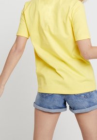 Żółta bawełniana koszulka z krótkim rękawem i luźnym krojem, zestawiona z niebieskimi dżinsowymi szortami z podwiniętymi nogawkami i tylnymi kieszeniami.