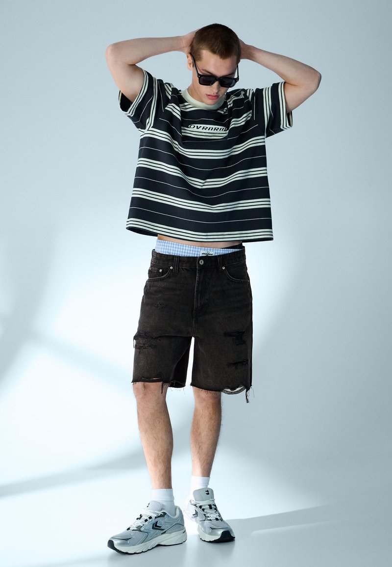 Jeune homme portant un t-shirt surdimensionné rayé noir et blanc, un short noir déchiré aux genoux, des baskets blanches et des lunettes de soleil noires, debout les mains sur la tête.