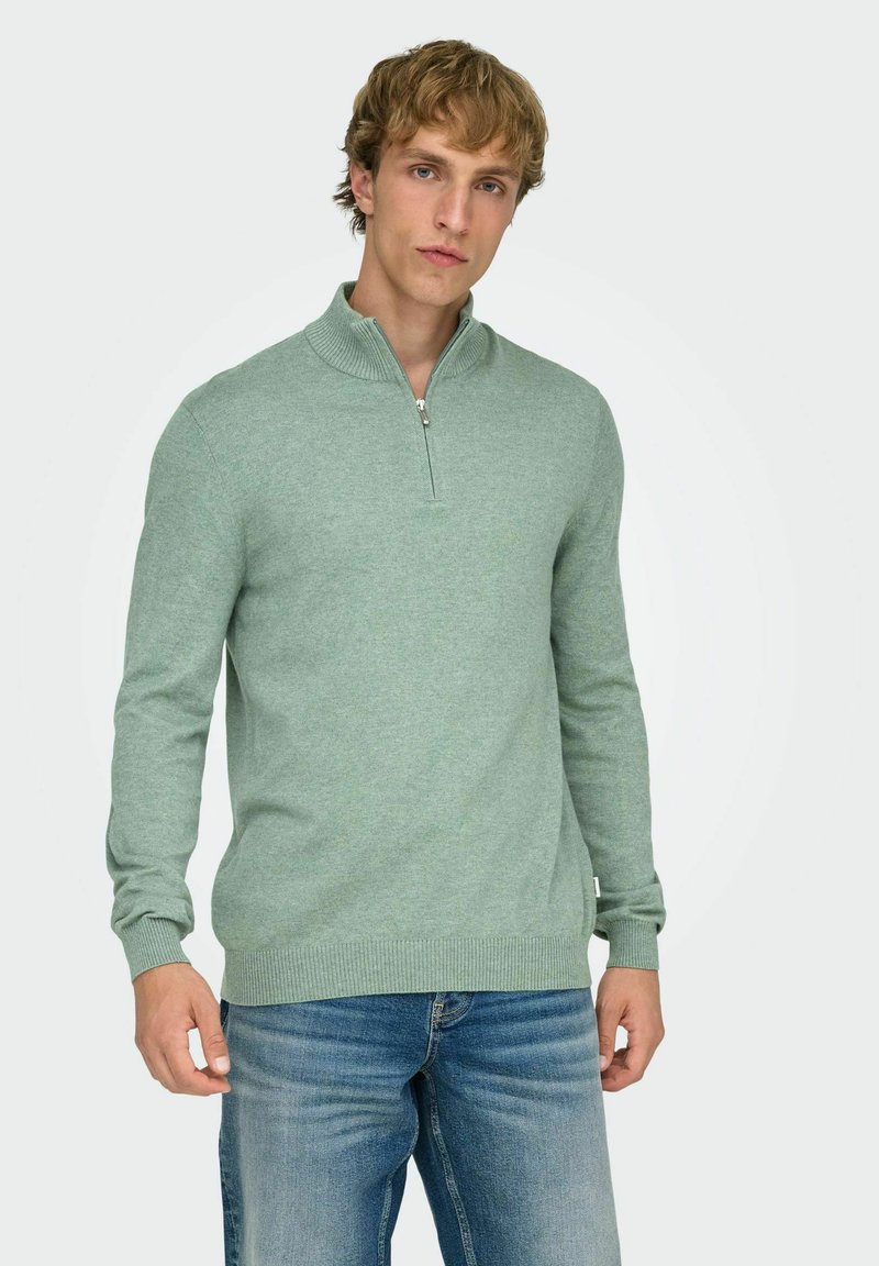 Mintgroene pullover trui met een ritssluiting aan de kraag, geribde manchetten en zoom. Zachte textuur, aangesloten pasvorm. Draagt bij een blauwe spijkerbroek.
