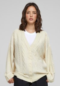Beige gestrickte Strickjacke mit einem Zopfmuster, V-Ausschnitt und großen Knöpfen. Lockere Passform mit gerippten Bündchen und Saum.