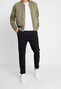 Olive zielona kurtka bomber z prążkowanym kołnierzem, zestawiona z czarnymi zwężanymi spodniami i białymi trampkami, wykonana z gładkiego materiału.