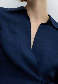 Námořnická modrá košile s límečkem, s výstřihem do V, přední kapsou a strukturovanou tkaninou. Čisté linie a střih přilehlý k tělu.