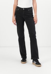 Persona che indossa jeans neri a gamba dritta, una maglietta bianca e sneakers nere con strisce bianche, in piedi contro uno sfondo bianco semplice.