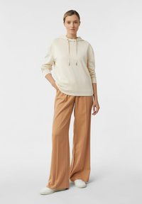 Felpa color beige chiaro con cappuccio e cordini, abbinata a pantaloni larghi color kaki. L'outfit presenta una vestibilità comoda e una texture di tessuto morbido.