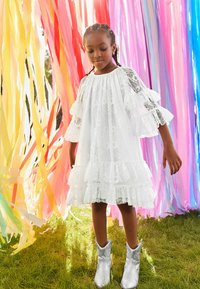 Robe en dentelle blanche avec des manches à volants et un ourlet en tiers, associée à des bottes de cowboy en argent brillant. L'arrière-plan présente des rubans colorés.