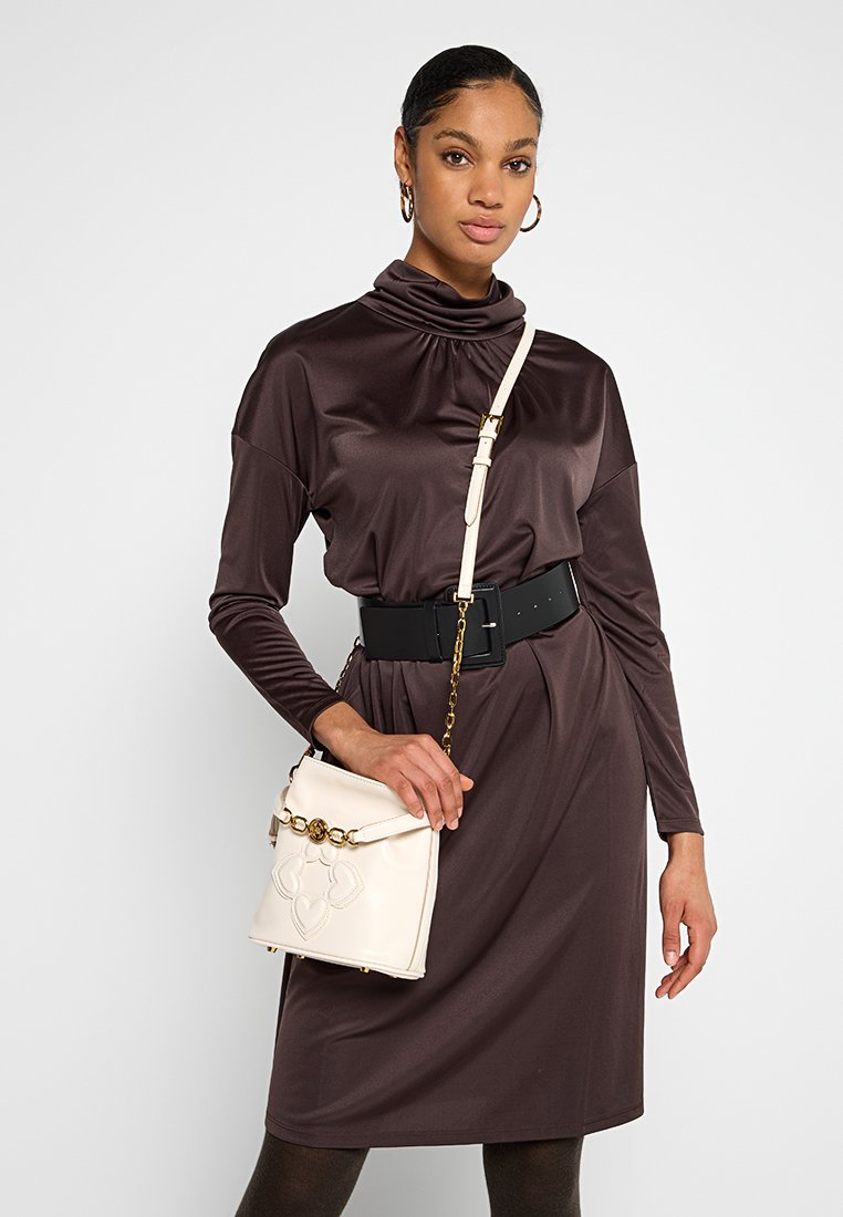 Frau in einem braunen Langarmkleid mit einem schwarzen Gürtel, die eine weiße Schultertasche mit goldenen Kettendetails hält.