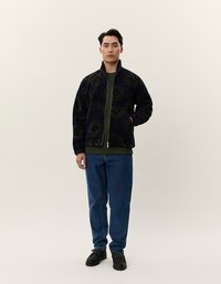 Marineblå fleecejakke med grønt mønster, glidelås foran, høy krage; lagd over grønn topp og kombinert med blå jeans og svarte sko.