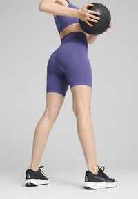 Pantaloni sportivi viola a vita alta con design testurizzato, abbinati a un crop top coordinato. La modella tiene in mano una palla medica nera e indossa scarpe da ginnastica nere.