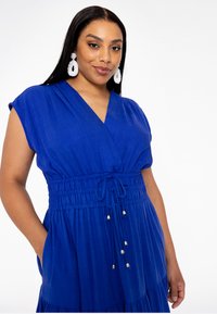 Blaues, kurzärmliges Kleid mit V-Ausschnitt und elastischem Bund. Hat ein gerafftes Oberteil und Taschen. Glatte Textur, leichter Stoff.