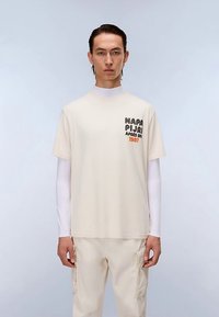 T-shirt in cotone color crema con maniche corte, con testo nero e arancione sul petto a sinistra. Indossata sopra uno strato a maniche lunghe bianco.