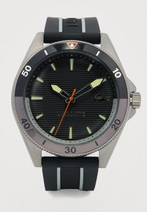 Zwarte en zilveren Lacoste horloge met gedurfde luminescente markeringen, oranje secondewijzer en een zwarte rubberen band met grijze strepen.