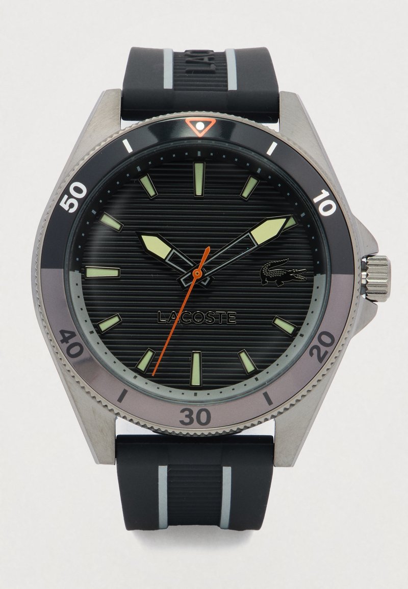 Zwarte en zilveren Lacoste horloge met gedurfde luminescente markeringen, oranje secondewijzer en een zwarte rubberen band met grijze strepen.