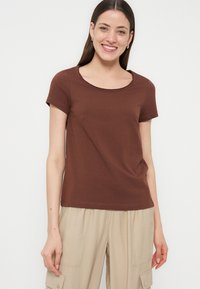 Γυναίκα με μακριά μαλλιά που χαμογελά φοράει καφέ κοντομάνικο πουκάμισο και μπεζ παντελόνι cargo σε απλό λευκό φόντο.