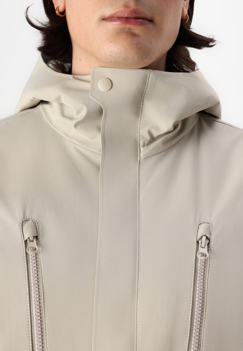 Chaqueta impermeable beige con un alto cuello, cierre a presión y dos bolsillos con cremallera que presentan cremalleras de color crema y una textura suave.