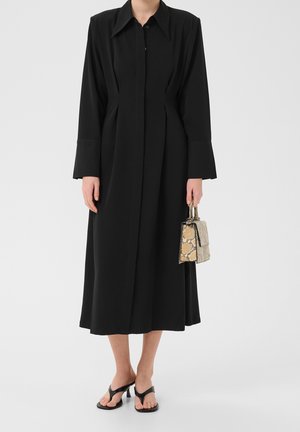 Personne portant un long manteau noir avec de larges poignets, tenant un petit sac à main en peau de serpent, et portant des sandales noires à talons ouverts.