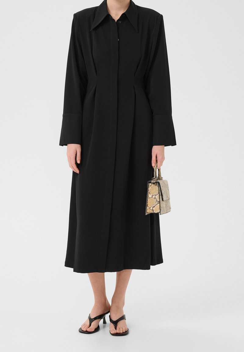 Personne portant un long manteau noir avec de larges poignets, tenant un petit sac à main en peau de serpent, et portant des sandales noires à talons ouverts.