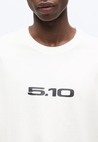 Κοντινό πλάνο ενός ατόμου που φοράει λευκό πουκάμισο με μαύρο κείμενο "5.10" στο κέντρο του στήθους, με απλό φόντο.