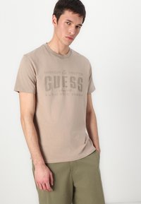 Béžové tričko s kulatým výstřihem a texturovaným povrchem, s velkým světle hnědým logem "GUESS" a dalším textem v mírně tmavším odstínu.