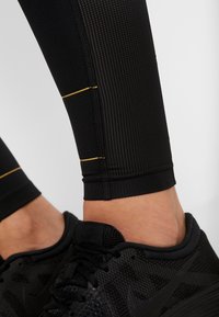 Legging sportive nere con un design testurizzato, caratterizzate da strisce verticali e dettagli gialli sul polpaccio, abbinate a scarpe da corsa nere.