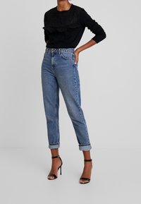 Haut noir à manches longues avec des volants texturés, associé à un jean bleu taille haute avec des ourlets retroussés et des talons aiguilles noirs à lanières.