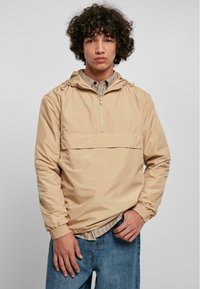 Beige letvægts pullover jakke med hætte, halv-lynlås fortil, elastiske manchetter og en frontlomme. Båret over en ternet skjorte.