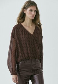 Frau trägt eine durchsichtige braune Bluse mit Pünktchen und langen Ärmeln sowie dunkelbraune Lederhose, steht vor einem neutralen Hintergrund.